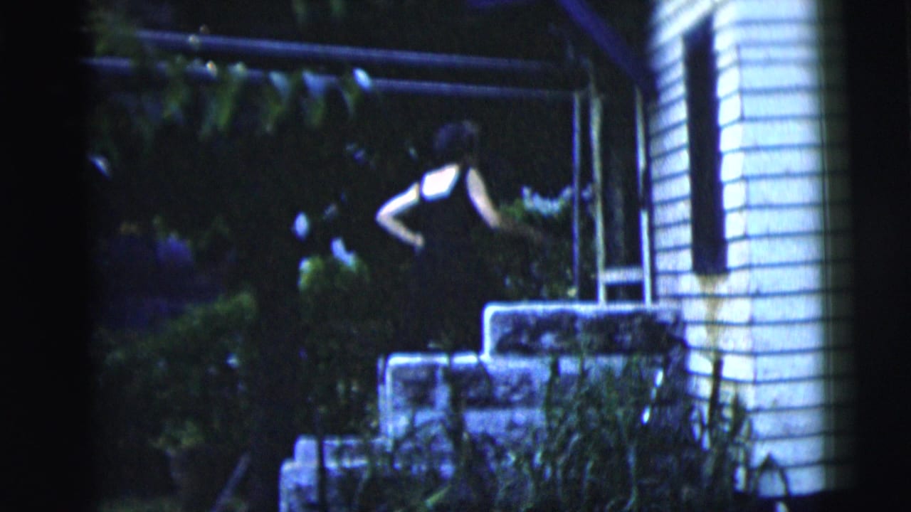 Woman in a black dress stands near stone steps beside a white-sided house at night.
