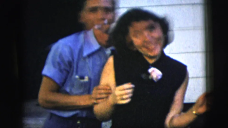 In 1958 New York City home-movie footage, a smiling woman in a black dress dances with a man in a blue shirt near house siding.