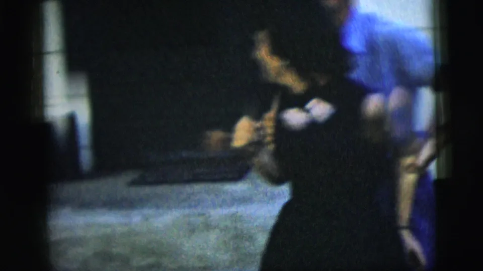 In 1958 New York City home-movie footage, a woman in a dark dress stands near a pale building wall while another adult moves behind her.