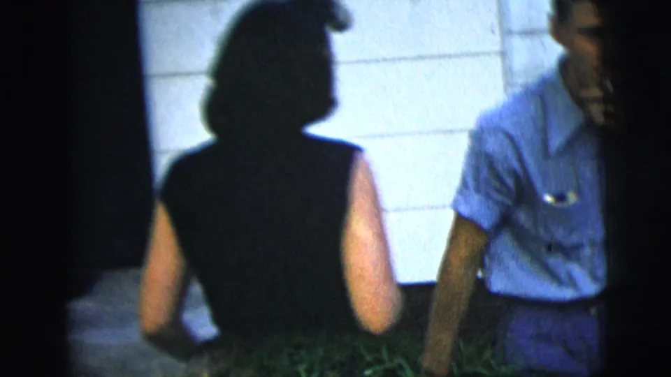 A woman in a sleeveless dark dress faces a man in a blue shirt near pale exterior walls and greenery in the New York City home movie.