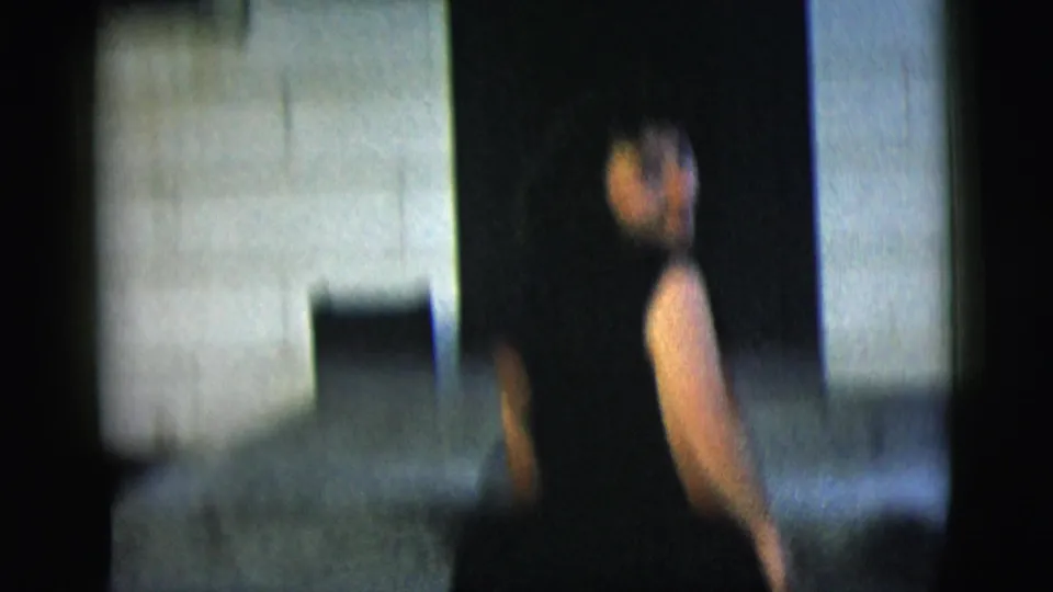 The home-movie camera follows a woman in a dark sleeveless dress as she moves near a pale building wall and patch of grass.