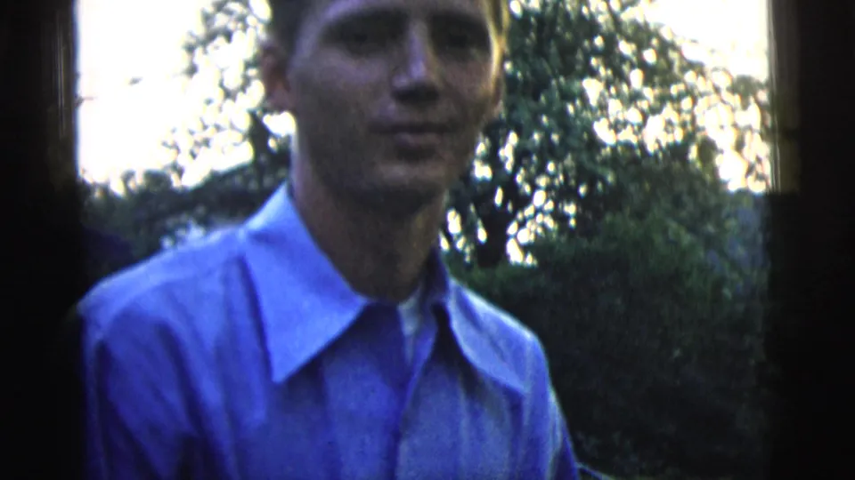 A man in a blue collared shirt faces the camera outdoors, with foliage behind him and the soft grain of 8mm footage visible.