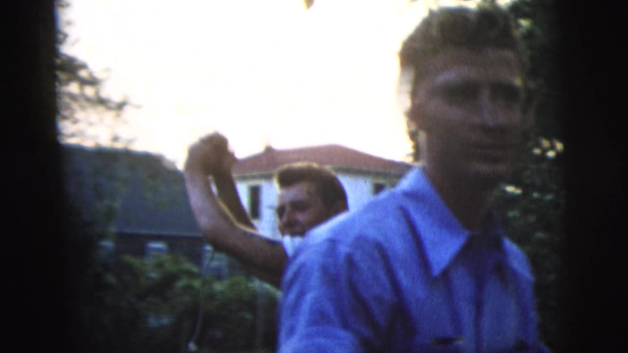 A man in a blue button-down shirt moves close to the camera as another young man works with a pole in a residential backyard.