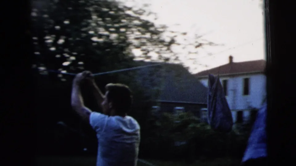 A young man in a white T-shirt holds a pole near a backyard clothesline, with houses and trees in the background.