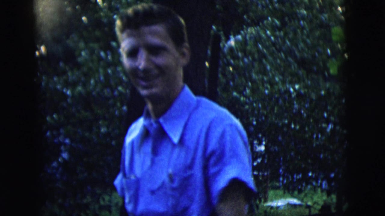 Young man in a blue short-sleeved shirt smiling in a leafy backyard.