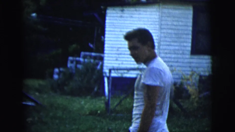 Adult man in a white t-shirt stands or walks in a backyard near a white-sided house in 1958 New York City footage.