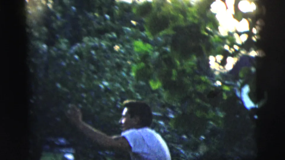 Adult man in a white t-shirt gestures in a leafy backyard, framed by trees and late-day light.