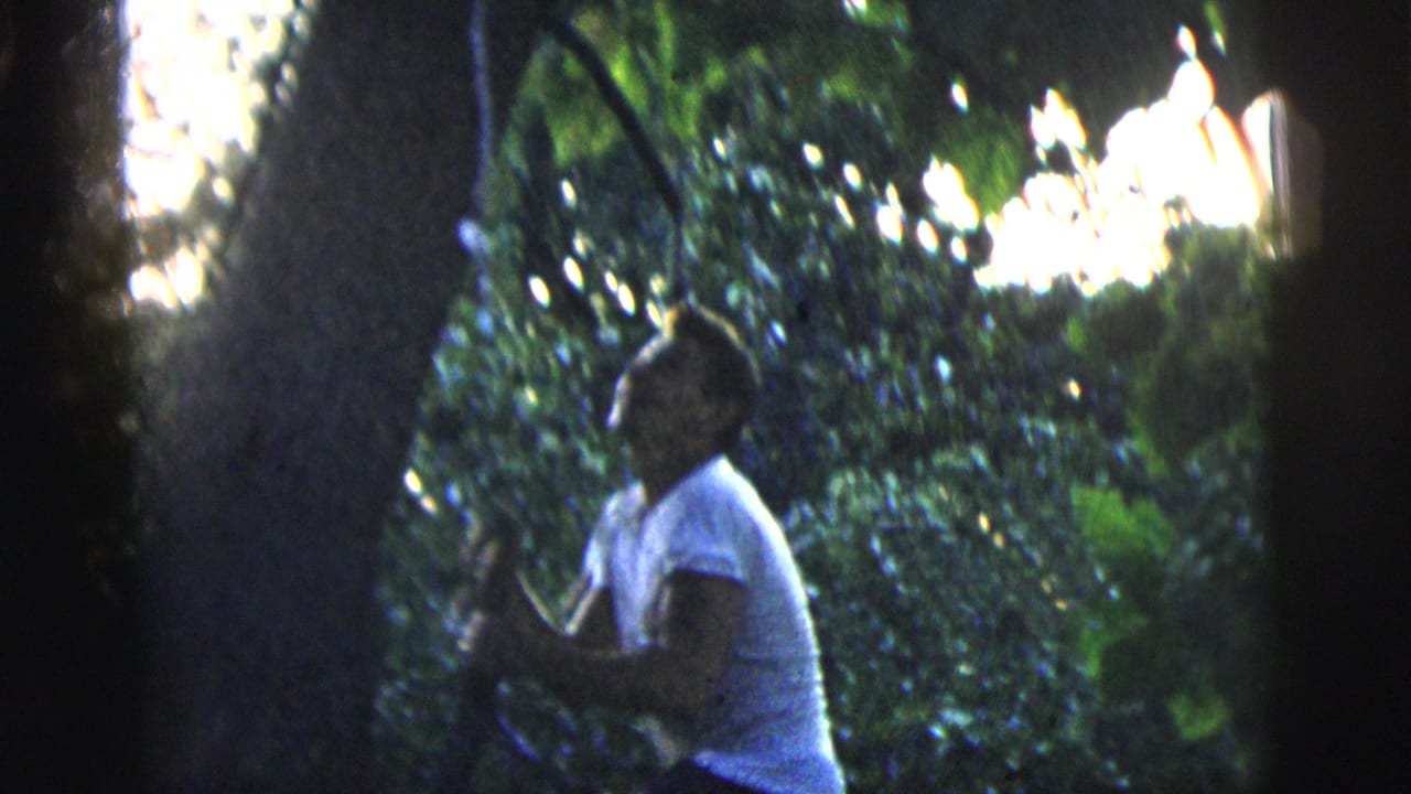 In 1958 New York City home-movie footage, a young man in a light T-shirt stands near a tree amid dark green foliage.