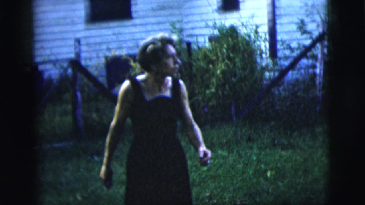 1958 New York City home-movie frame of a woman in a dark sleeveless dress walking in a grassy backyard near a house.