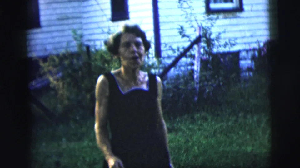 1958 New York City 8mm frame showing the woman closer to camera in the backyard, with house siding and fencing behind her.