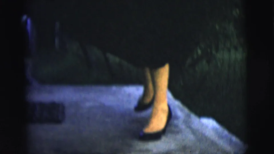 Adult woman in a dark dress and heels steps along a backyard path in 1958 New York City home-movie footage.