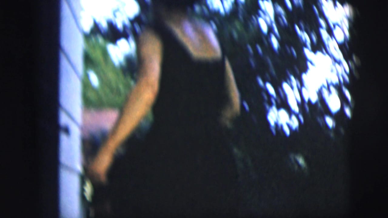 Woman in a dark dress stands near a house and trees in soft-focus home-movie footage.