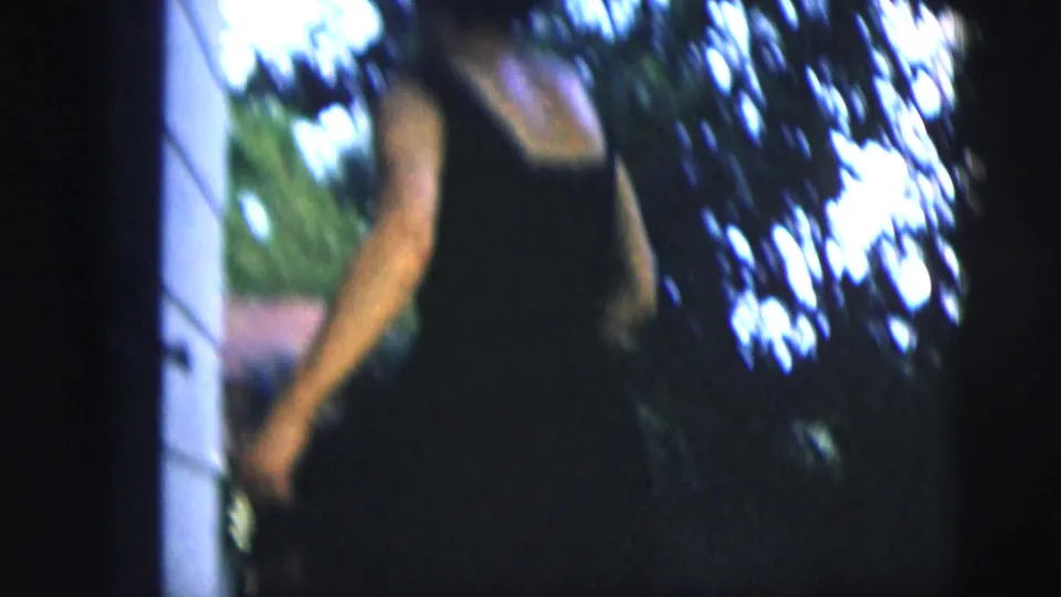 Soft 1958 8mm frame of a woman in a dark sleeveless dress near a house exterior and leafy trees in New York City metadata context.
