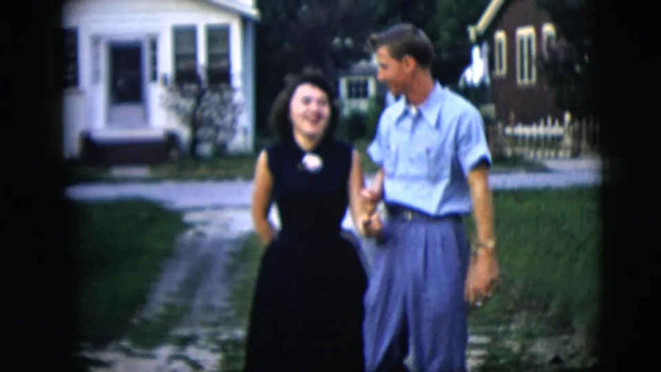 A smiling couple walks arm-in-arm in a New York City 1958 home-movie frame, with a small porch, lawn, and neighboring houses behind them.
