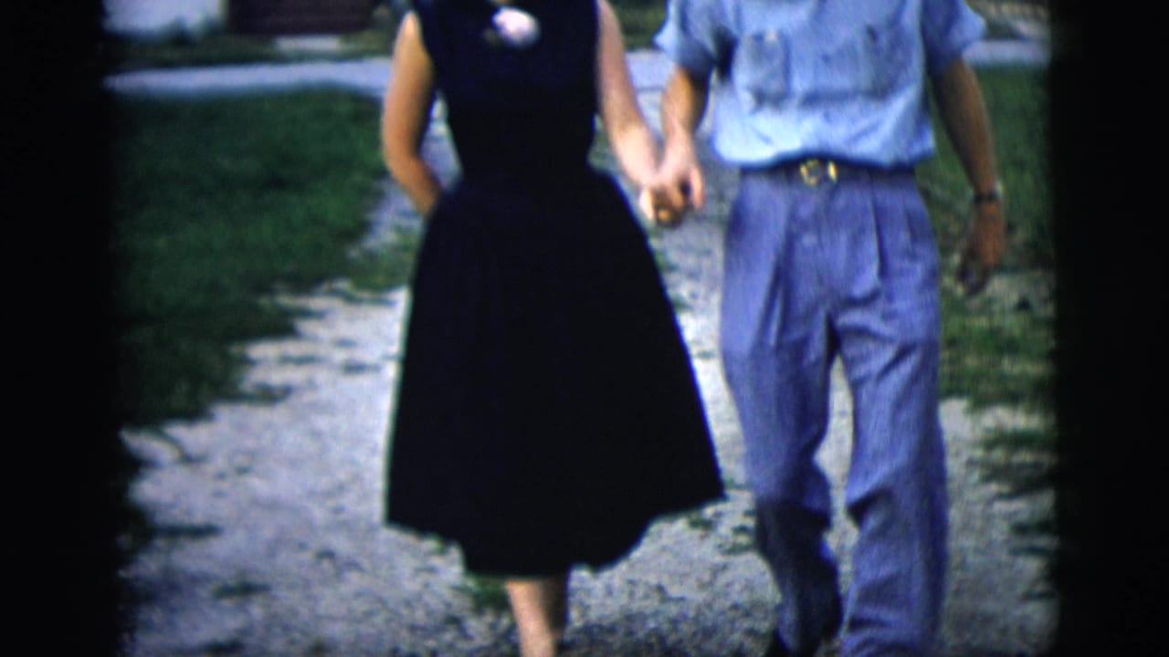 The couple's clasped hands and mid-century clothing are visible as they walk along a gravel driveway in the residential scene.