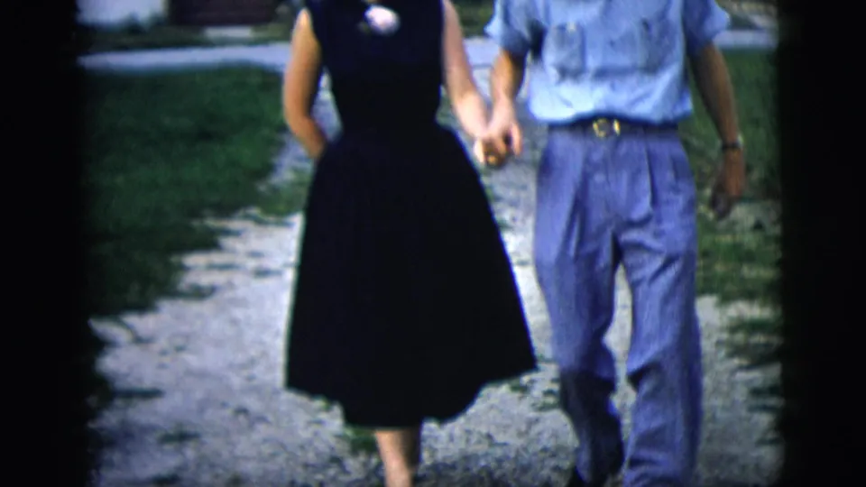 The couple's clasped hands and mid-century clothing are visible as they walk along a gravel driveway in the residential scene.