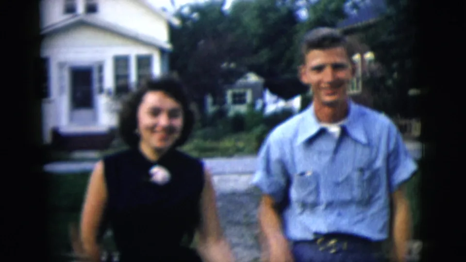 The woman and man face the camera with smiles, with a white house, lawn, and neighborhood walkway visible behind them.