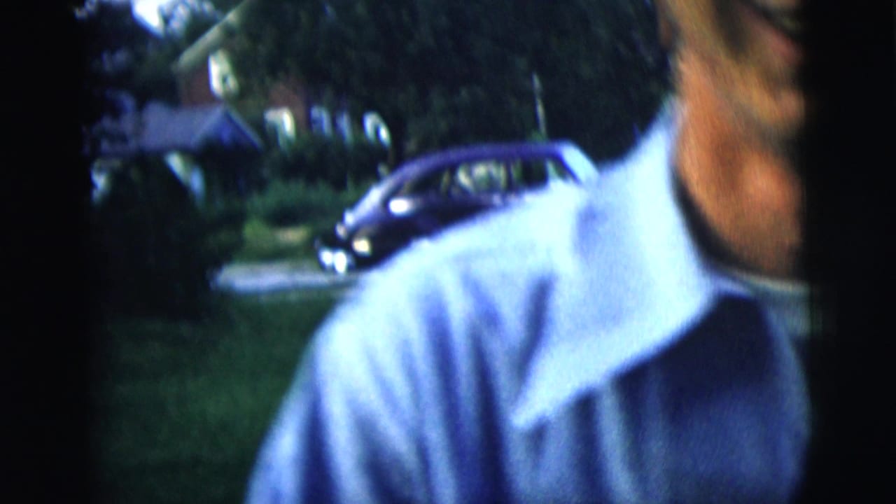 The camera briefly crops close on the man in a light blue shirt, with a lawn, trees, and parked vintage car behind him.