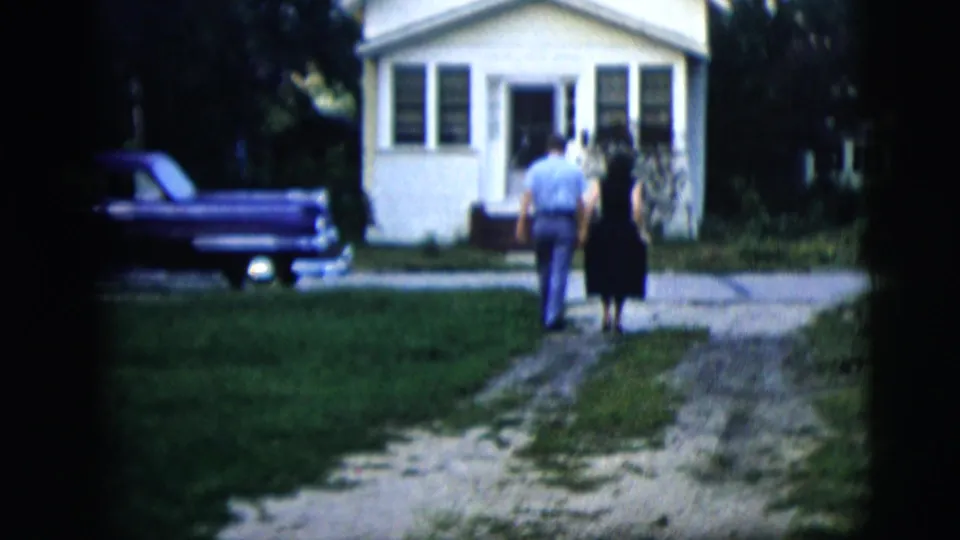 The couple walks away from the camera toward a small suburban house, holding hands along a gravel path beside a purple car.