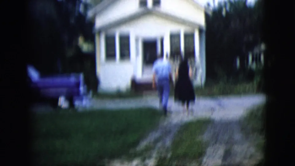 In a soft 8mm frame, the couple nears the front entrance of the small house, with lawn, trees, and parked car still visible.