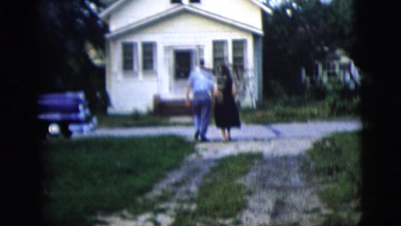 Two adults walk hand-in-hand up a gravel driveway toward a small white house.