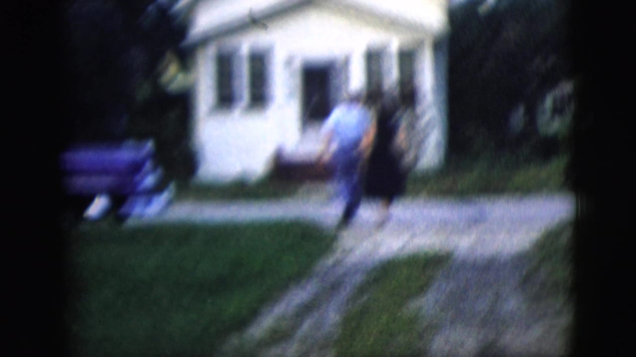 The couple continues toward the white house in a soft, motion-blurred 8mm frame with lawn and parked car visible.
