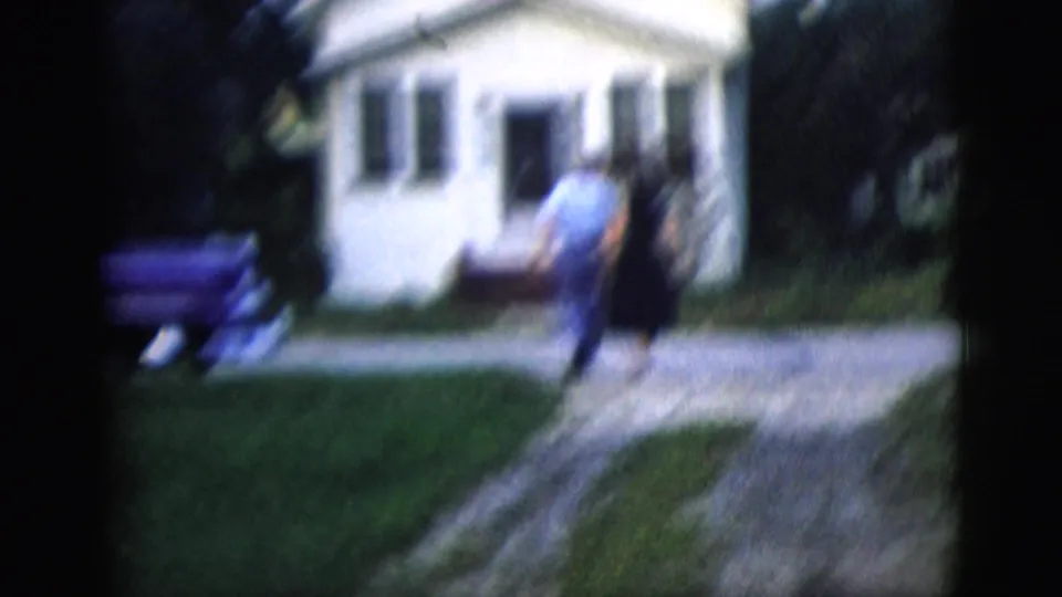 The couple continues toward the white house in a soft, motion-blurred 8mm frame with lawn and parked car visible.