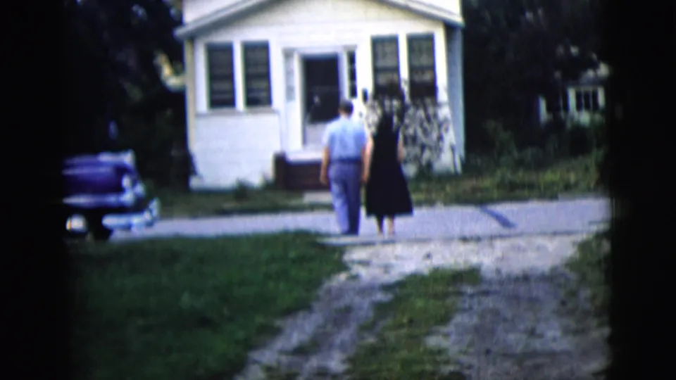 Two adults appear closer to the house, with the front doorway, windows, driveway, lawn, and parked car in view.