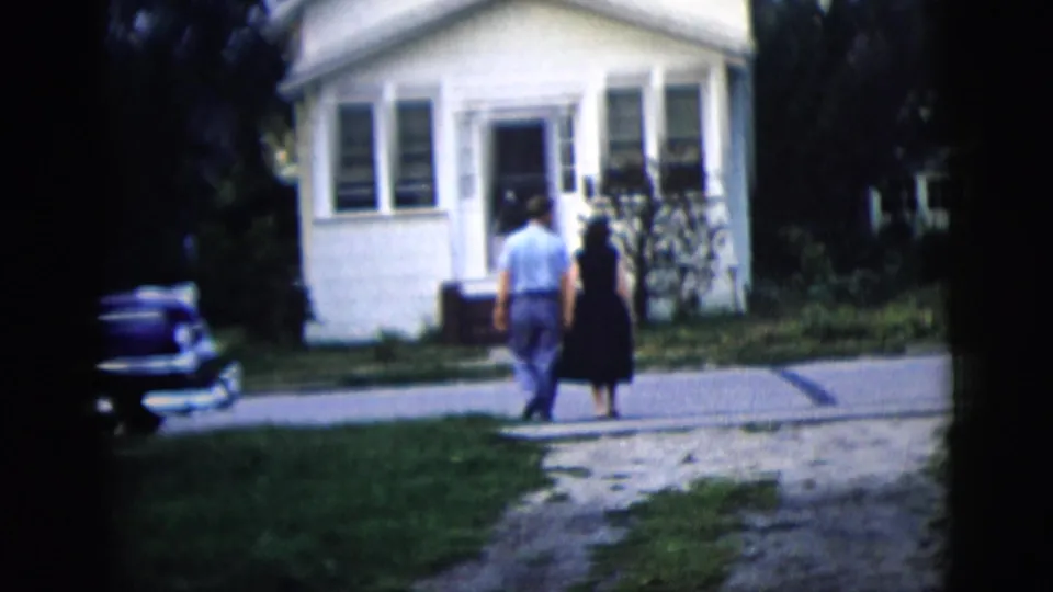 The couple is seen from behind near the driveway and street edge, with the small white house and parked car behind them.