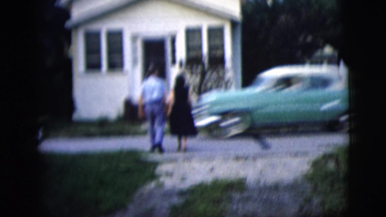 1958 home-movie frame of a couple walking toward a small white house in New York City with a turquoise 1950s car by the curb.