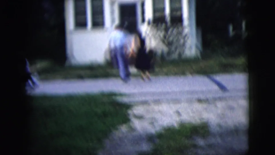 1958 home-movie frame of a couple walking from the street toward a small white house, seen from behind in soft focus.