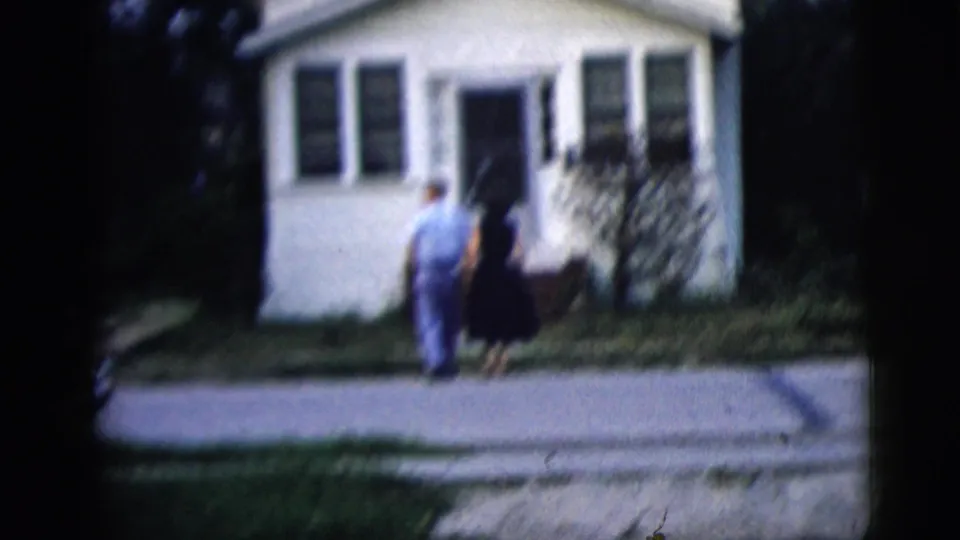 1958 New York City home-movie frame of a couple near the sidewalk in front of a small white house with shrubs and windows.