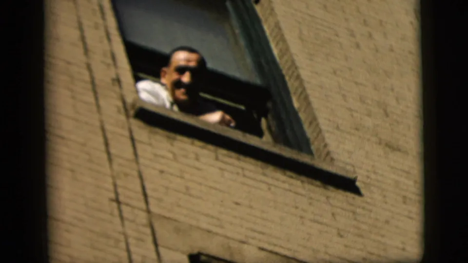 A man in a white shirt smiles from an open window on a brick building in 1958 New York City footage.