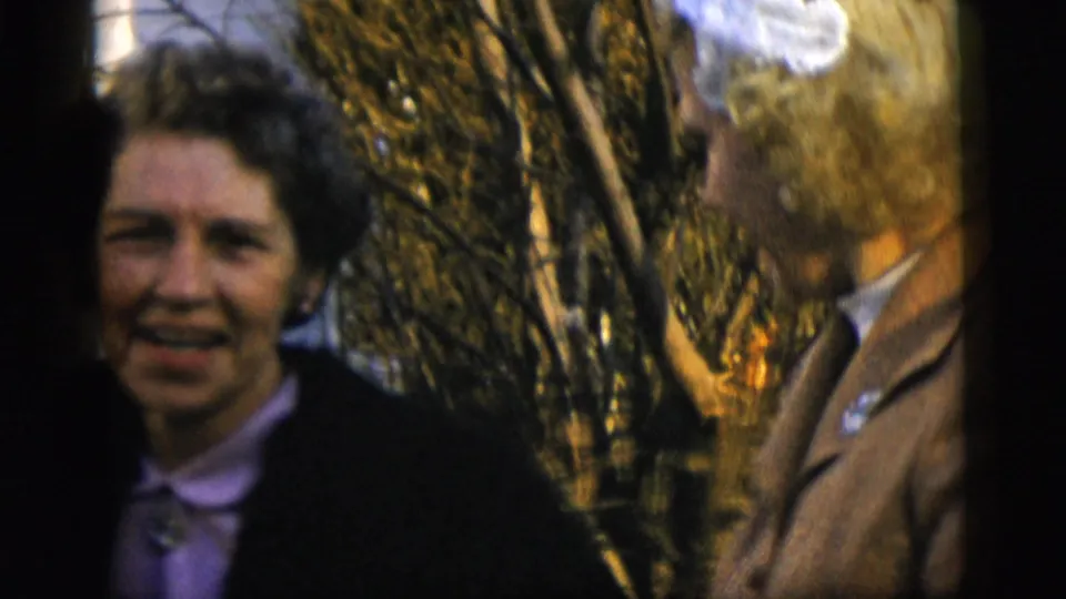 A woman with short dark hair faces the camera near another adult, with bare branches behind them in 1958 New York City footage.