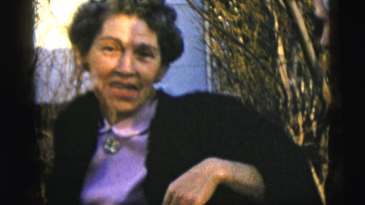 In 1958 New York City home-movie footage, an adult woman in a dark coat and purple blouse faces the camera outdoors by bare shrubs.