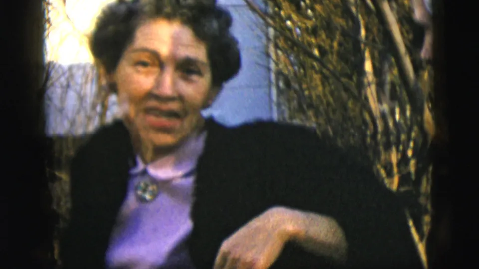 In 1958 New York City home-movie footage, an adult woman in a dark coat and purple blouse faces the camera outdoors by bare shrubs.