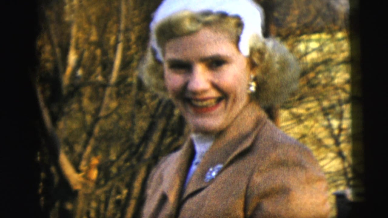 Smiling blonde adult woman wearing a white hat and brown suit outdoors near bare branches.