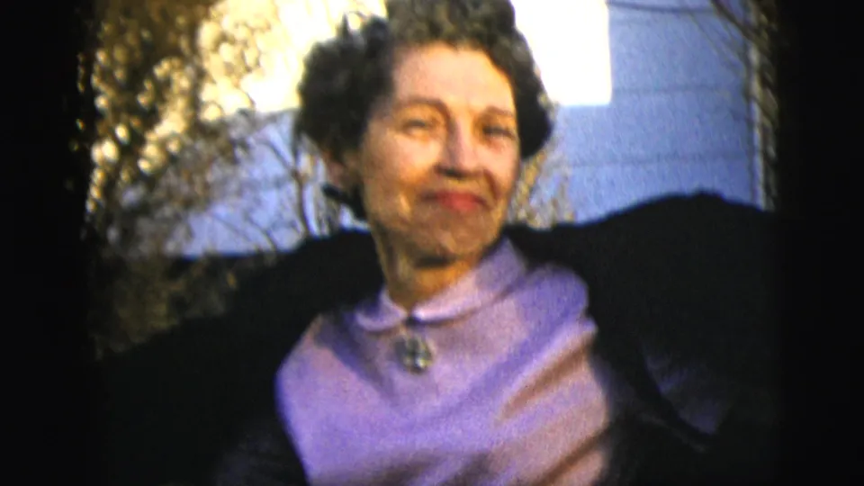 The dark-haired woman in a dark coat and purple blouse appears in soft 8mm focus outdoors, framed close against shrubs and sky.