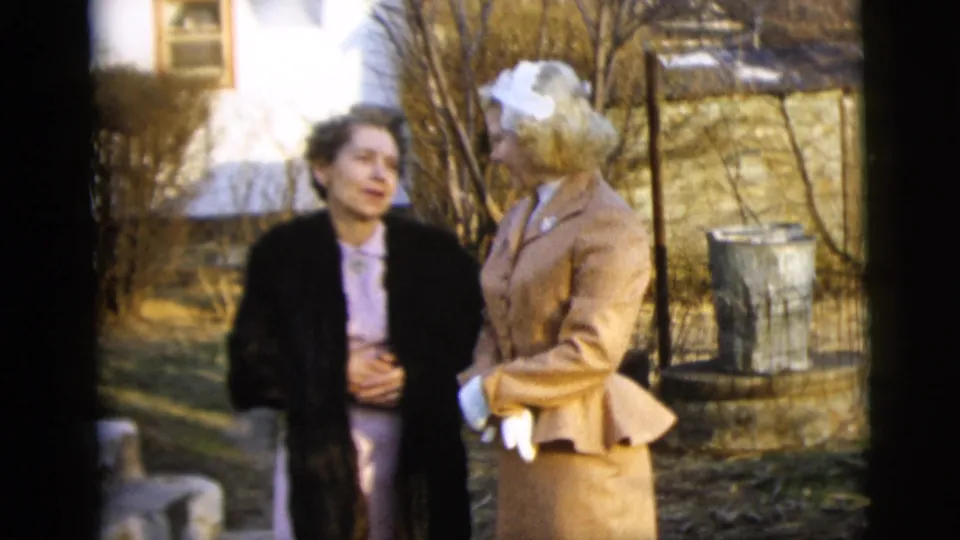 Two adult women stand in conversation in a New York City yard, with bare trees, a house, and a metal bin visible behind them.