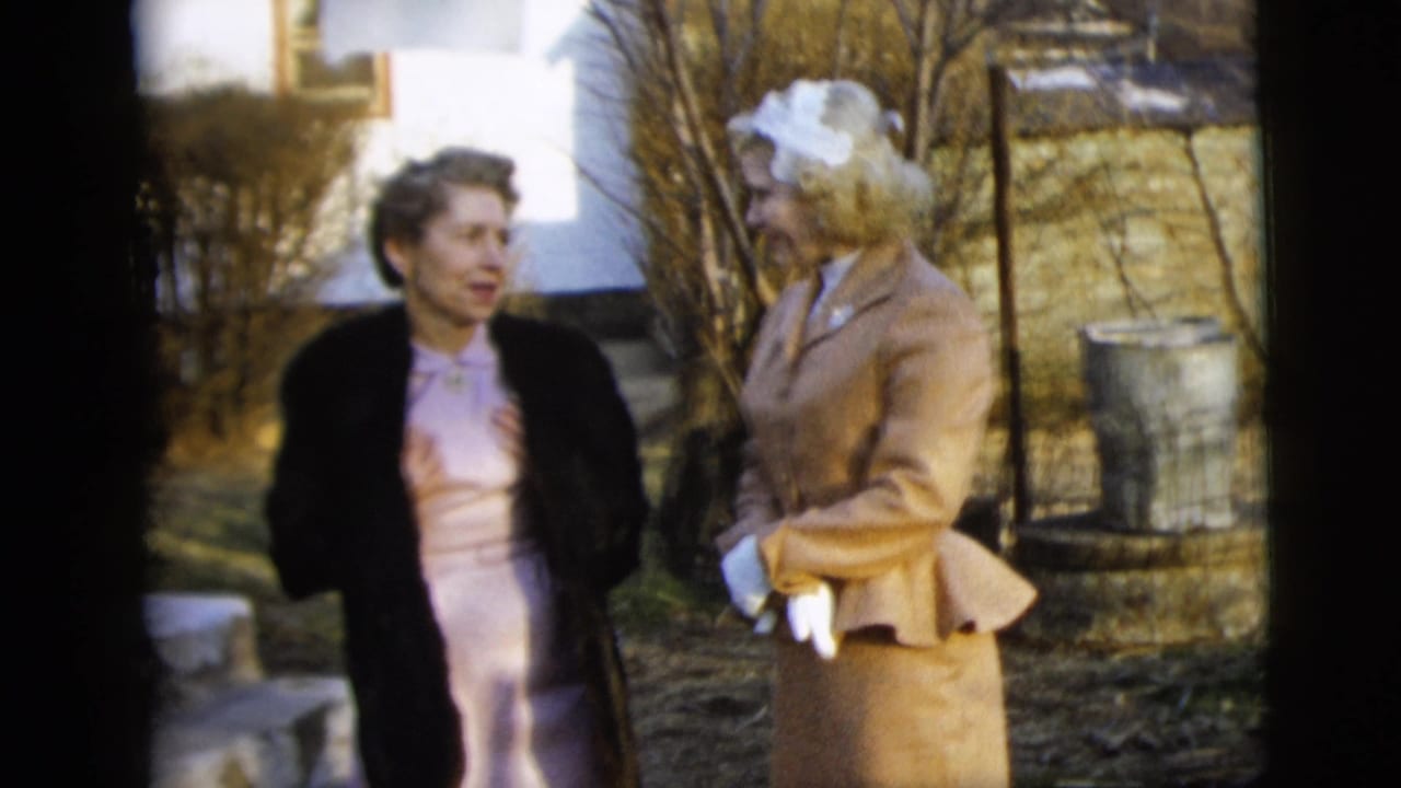 1958 New York City home-movie frame of two women outdoors, one in a pale dress and dark fur coat and the other in a tan suit with white gloves and hat.
