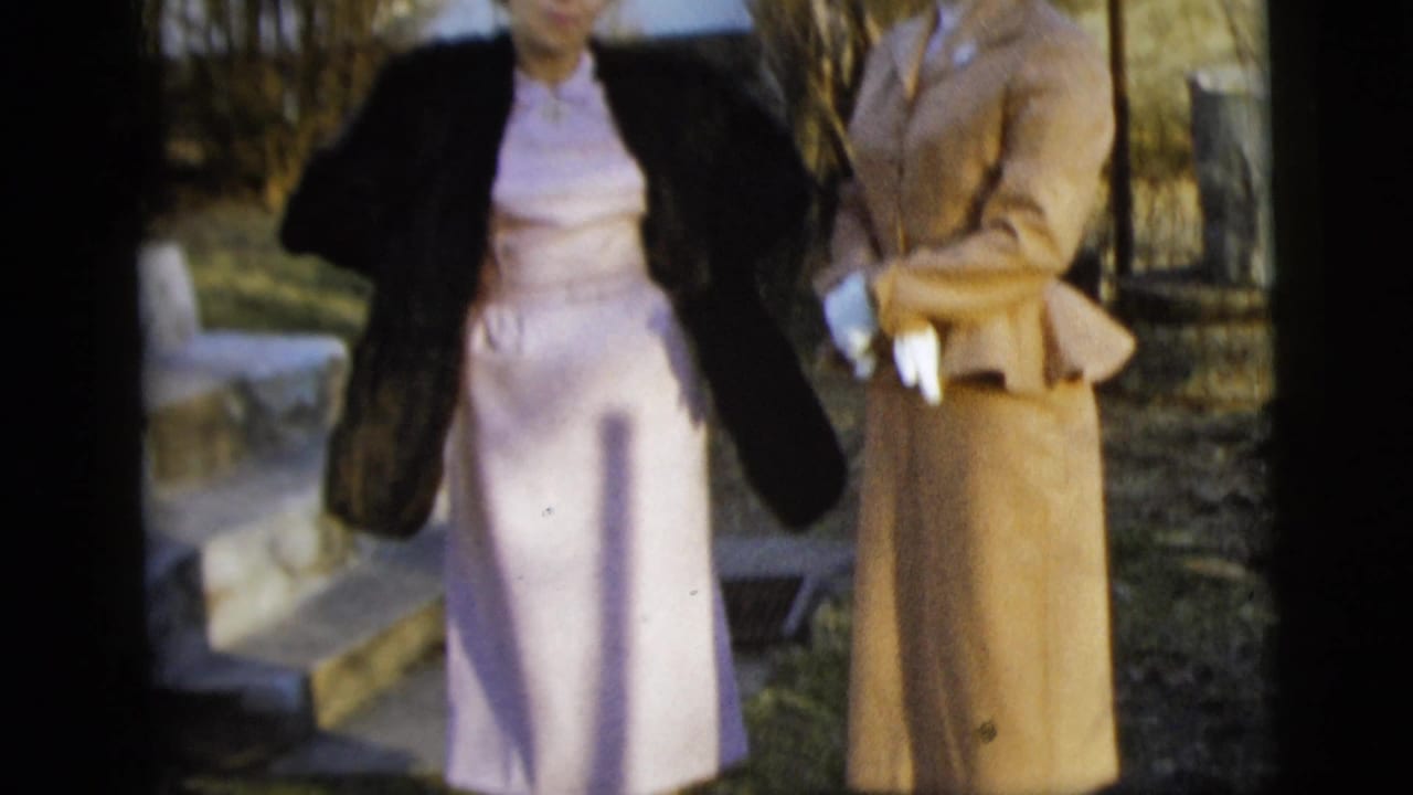 Cropped 1958 New York City home-movie frame showing two women in formal dress, including a dark coat, pale dress, tan suit, and white gloves.