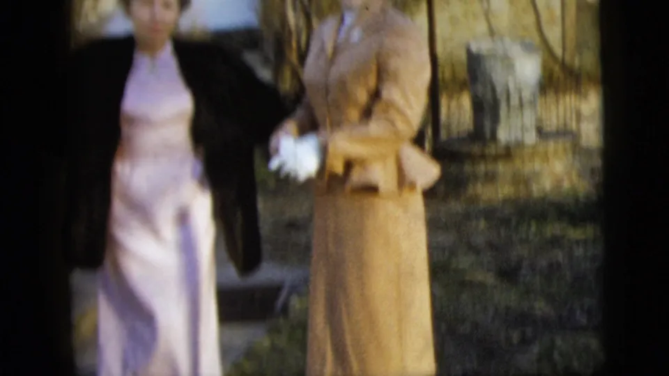 Close cropped 1958 New York City home-movie frame of two women outdoors, with one in a pale dress and dark coat and the other in a tan suit holding white gloves.