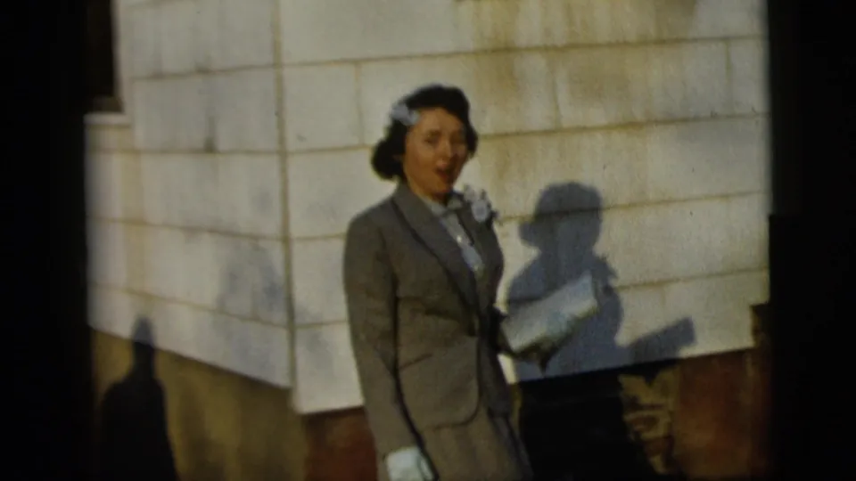 1958 New York City home-movie frame of a woman in a gray suit and white gloves standing by a light exterior wall, her face turned as if speaking or reacting.