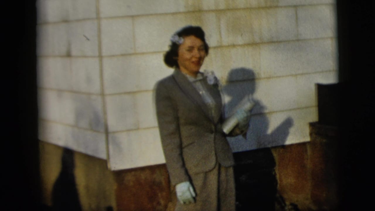 In 1958 New York City, a woman wearing a grey suit, white gloves, corsage, and hair bow poses beside a tiled wall.
