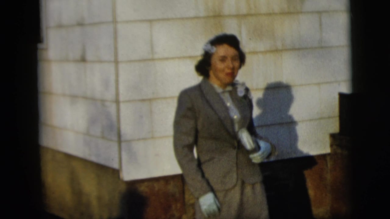 The woman remains posed beside the wall, with gloves, corsage, and a rolled document or program visible in the sunlight.