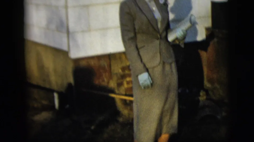 A cropped 8mm frame shows the woman's formal suit, gloved hand, corsage area, tiled wall, and sunlit shadows.