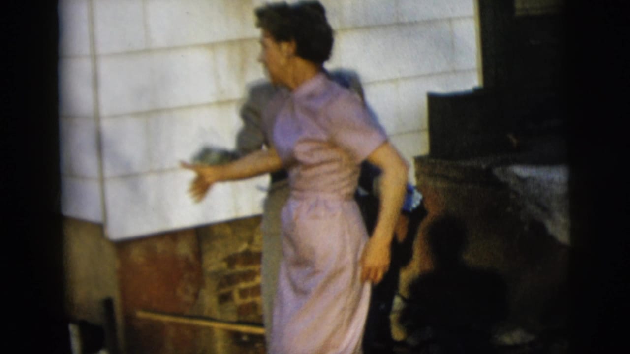 In 1958 New York City home-movie footage, a woman in a lavender dress turns beside white siding and brickwork in bright sun.