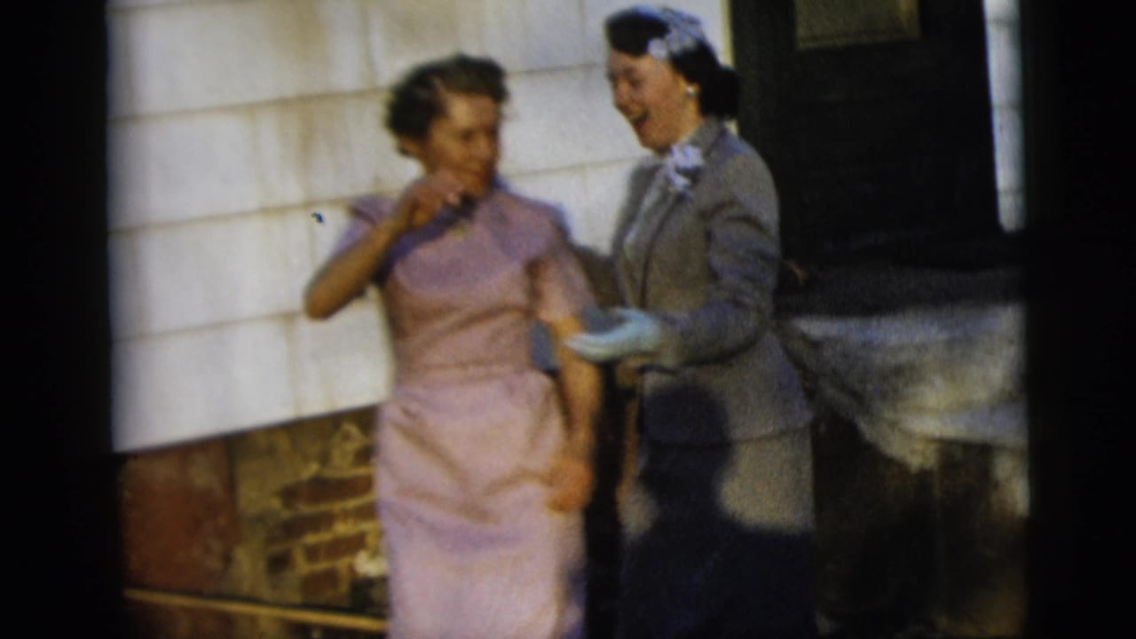 Two women in 1950s attire, one in a lavender dress and one in a gray suit with a head covering, smile outdoors in New York City.