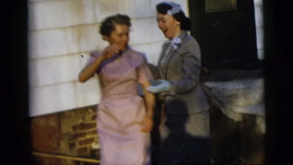 Two women in 1950s attire, one in a lavender dress and one in a gray suit with a head covering, smile outdoors in New York City.