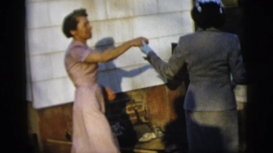 The women face each other in a playful dance gesture, with white siding, brickwork, and hard sunlight behind them.